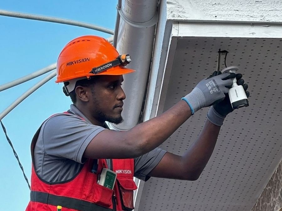 A technician in an orange Hikvision helmet fitting a CCTV camera under a roof edge